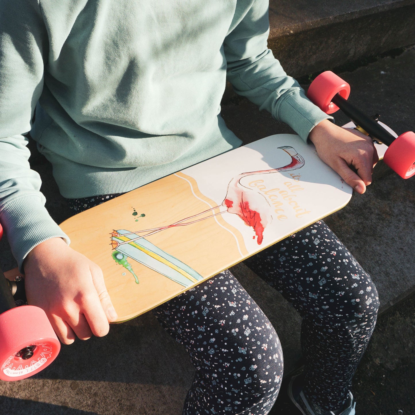 Ein Mädchen hält das BTFL Kinder Longboard mit Flamingo Design auf dem Schoss