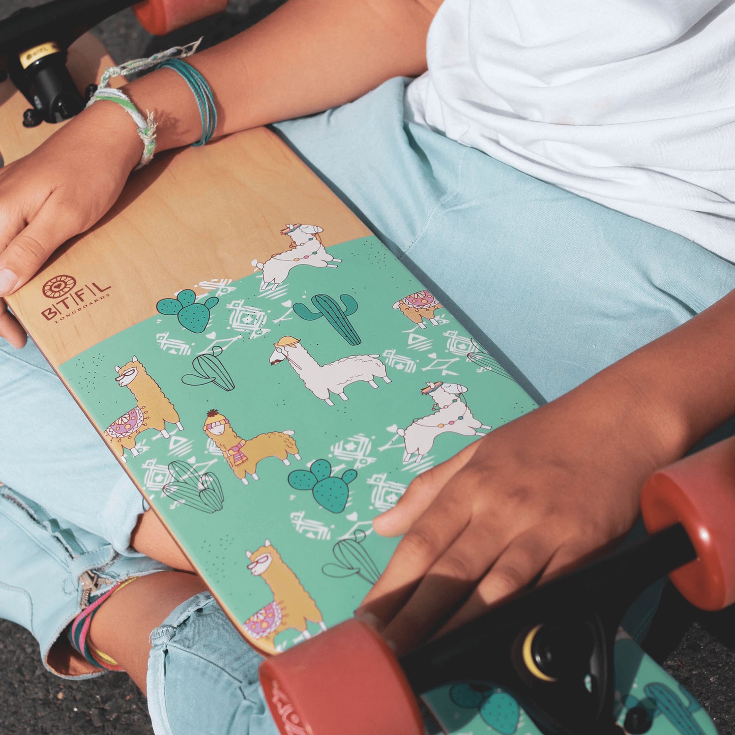 Ein Mädchen hält ein Longboard für Kinder auf dem Schoss, darauf sind kleine Lamas abgebildet