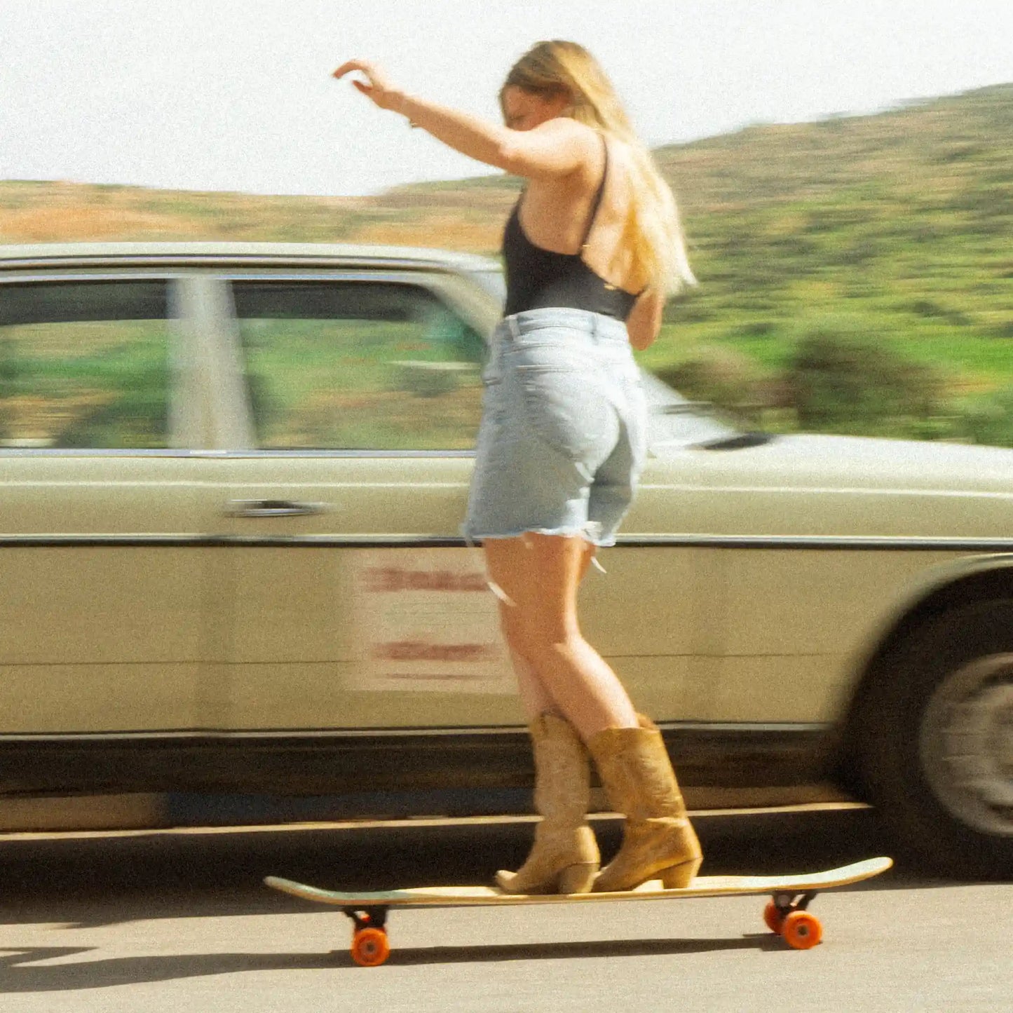 Eine Frau macht einen sogenannten Cross Step auf dem Longboard, der Hintergrund ist verschwommen, man sieht ein Auto und Bäume